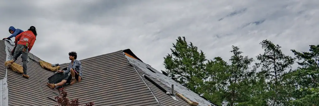 Three roofers high on a brick house mid re-roof: one in a blue hood at the ridge, one in a red shirt hauling felt across the slope, one sat on the bare deck with a shingle bundle, under a heavy grey sky.