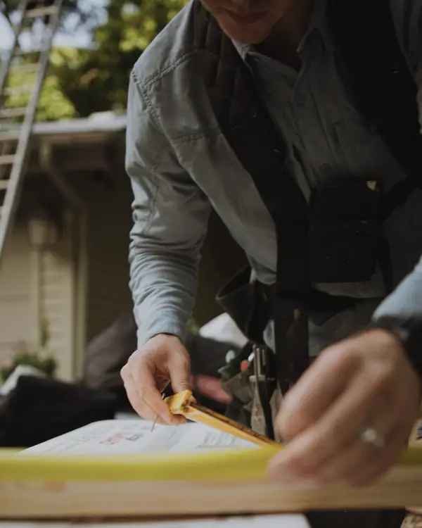 Close on a tradesman's hands marking a wrapped bundle with a carpenter's pencil, a yellow folding rule held along the edge, tool-belt straps across his work shirt and a ladder against the house behind him.