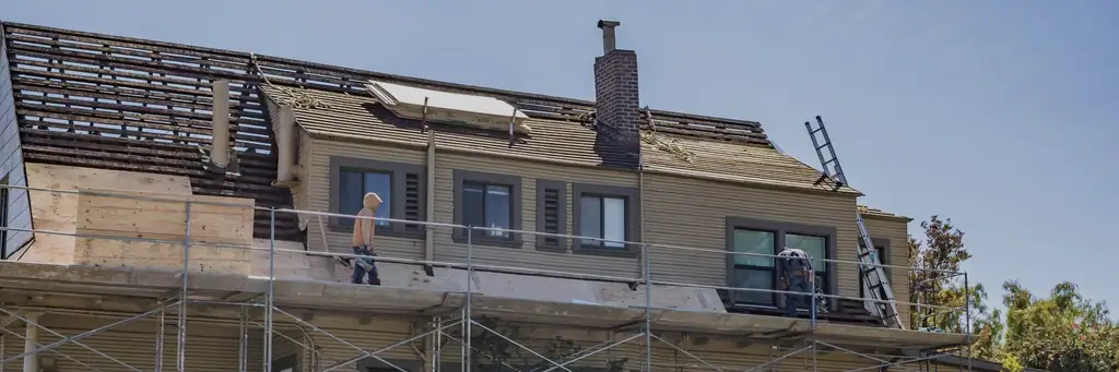 A timber house with its roof taken back to bare rafters, scaffolding across the front, a worker walking the staging plank and a second at an upper window.