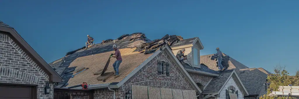 A stone-and-brick house at golden hour with the roof decking stripped bare, five crew working across it and torn shingles sliding down the slope against a deep blue sky.