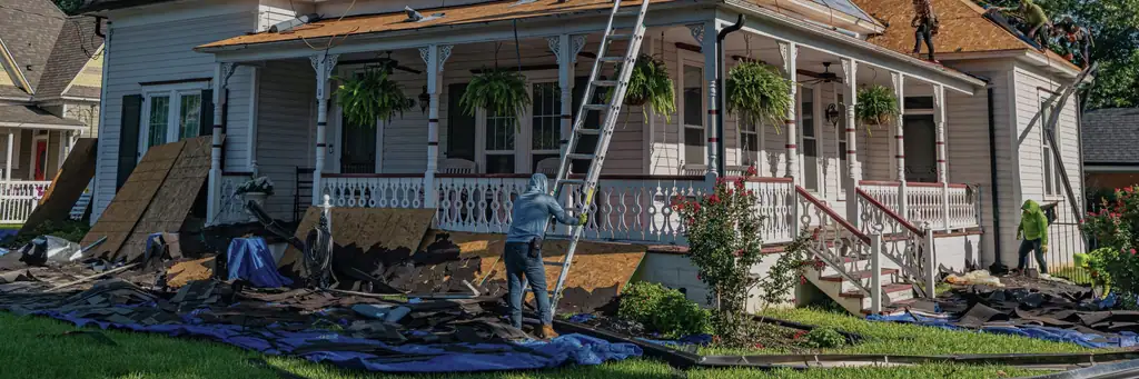 The front lawn of a lived-in white Victorian house with hanging ferns on its porch, an extension ladder to the eave, a crew member dragging torn-off shingles across the grass and more of the crew working the bare deck above.