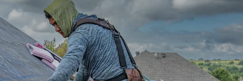 A roofer in a hooded sun-shirt bent to the work on a shingle slope under a heavy grey sky, bundles staged on the deck behind him and the neighbouring rooftops beyond.